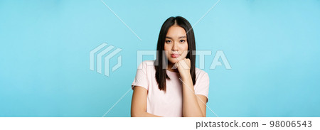 Asian girl watching smth boring, looking with unamused, reluctant face expression, standing in t-shirt over blue backround 98006543