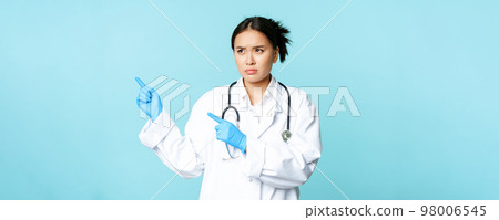 Angry asian medical worker, nurse or doctor express disapproval, pointing left and furrow eyebrows, standing over blue background 98006545
