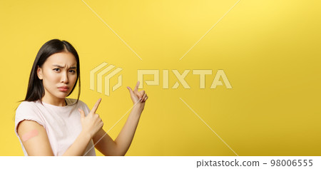 Covid-19 vaccination concept. Disappointed sulking asian girl pointing at promo banner, being vaccinated from coronavirus, yellow background Covid-19 vaccination concept. Disappointed sulking asian girl pointing at promo banner, being vaccinated from coronavirus, yellow background 98006555