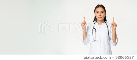 Smiling asian female physician, pointing fingers up and looking at camera, provide medical healthcare information, white background Smiling asian female physician, pointing fingers up and looking at camera, provide medical healthcare information, white background 98006557