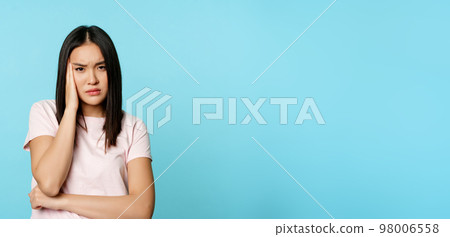 Troubled young asian woman holding hand on head, frowning and staring with displeased, concerned face, standing in t-shirt over blue background Troubled young asian woman holding hand on head, frowning and staring with displeased, concerned face, standing in t-shirt over blue background 98006558