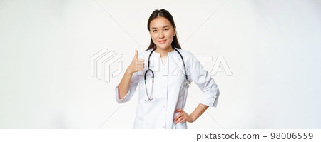 Smiling professional doctor in medical uniform shows thumbs up. Pleased asian female physician confirms smth, recommending product, white background Smiling professional doctor in medical uniform shows thumbs up. Pleased asian female physician confirms smth, recommending product, white background 98006559