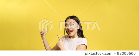 Covid-19 vaccination concept. Happy vaccinated korean girl with patch on arm, pointing at promo banner, showing advertisement and smiling Covid-19 vaccination concept. Happy vaccinated korean girl with patch on arm, pointing at promo banner, showing advertisement and smiling 98006606