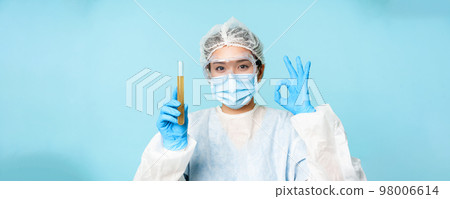 Laboratory worker, nurse in personal protective equipment, showing sample in test tube and okay sign, testing patient analise, blue background 98006614