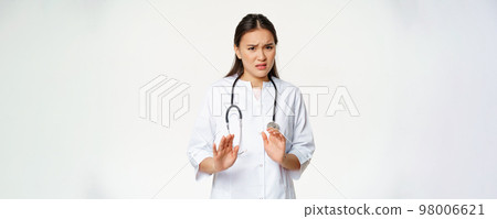 Disgusted asian female doctor rejecting, saying no, looking with dislike, refusing from smth unpleasant, standing in medical uniform against white background Disgusted asian female doctor rejecting, saying no, looking with dislike, refusing from smth unpleasant, standing in medical uniform against white background 98006621