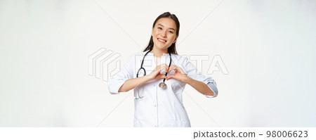 Smiling asian female doctor care for her patients, shows heart gesture and looking happy, standing in medical clinic uniform, white background Smiling asian female doctor care for her patients, shows heart gesture and looking happy, standing in medical clinic uniform, white background 98006623
