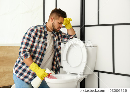Young man feeling disgust while cleaning toilet bowl in bathroom 98006816