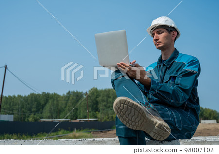 Caucasian male builder in hardhat sits on floor slabs and uses laptop at construction site. Caucasian male builder in hardhat sits on floor slabs and uses laptop at construction site. 98007102
