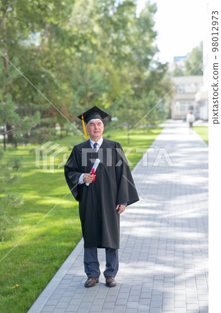 An old man in a graduation gown walks outdoors and holds a diploma. 98012973