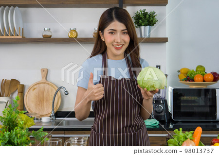 Young asian housewife dressed in an apron raise finger thumb up while holding fresh lettuce. Morning atmosphere in a modern kitchen. Young asian housewife dressed in an apron raise finger thumb up while holding fresh lettuce. Morning atmosphere in a modern kitchen. 98013737