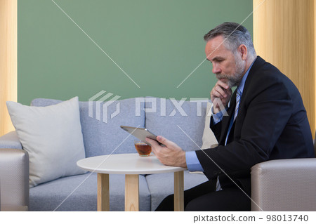 Senior businessman in suit and necktie reading email on tablet computer in cafeteria during coffee break. A cup of hot tea are on the table. 98013740
