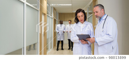 Senior professor consults a female doctor in front of a hospital patient's room. Both of them pay attention to the document on the clipboard. Two doctors walking on corridor in the background. 98013899