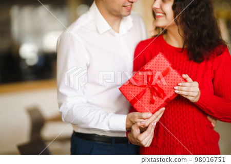 Man gives to his woman a gift box with red ribbon. A loving couple cuddles. Man gives to his woman a gift box with red ribbon. A loving couple cuddles. 98016751