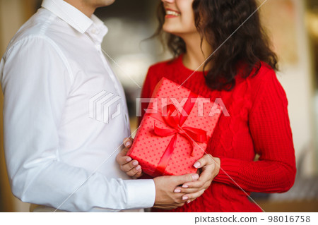 Man gives to his woman a gift box with red ribbon. A loving couple cuddles. Man gives to his woman a gift box with red ribbon. A loving couple cuddles. 98016758