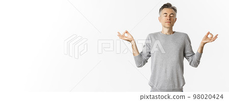 Portrait of calm and relaxed middle-aged man, breathing air freely, meditating with eyes closed and hands spread sideways, practice yoga over white background 98020424