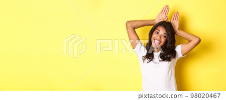 Portrait of attractive african-american girl, smiling and showing bunny ears on head, standing silly and coquettish over yellow background 98020467
