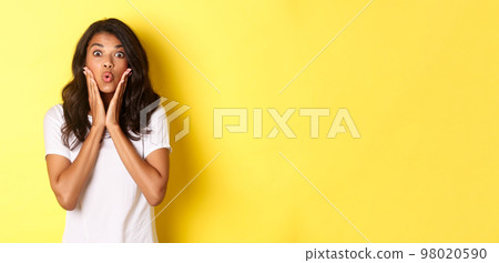 Portrait of attractive african-american teenage girl looking amazed, saying wow and stare at camera excited, standing over white background 98020590