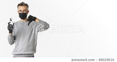 Concept of coronavirus, lifestyle and quarantine. Portrait of middle-aged man in medical mask and gloves do not recommend antiseptic, showing hand sanitizer and thumbs-down, white background Concept of coronavirus, lifestyle and quarantine. Portrait of middle-aged man in medical mask and gloves do not recommend antiseptic, showing hand sanitizer and thumbs-down, white background 98020610