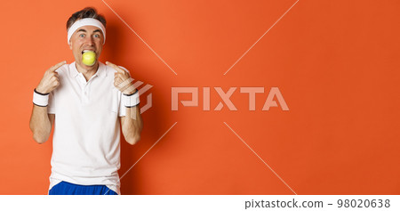 Portrait of funny, handsome middle-aged fitness guy, holding apple with teeth and pointing at it, eating healthy and working out, standing over orange background 98020638