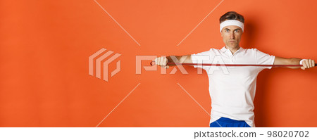 Concept of workout, gym and lifestyle. Serious and confident middle-aged fitness guy, holding stretching rope for exercises, standing over orange background 98020702