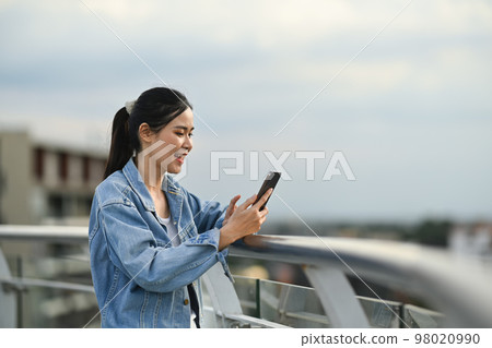 Happy young woman using smart phone while standing on terrace against background of beautiful evening sky background 98020990