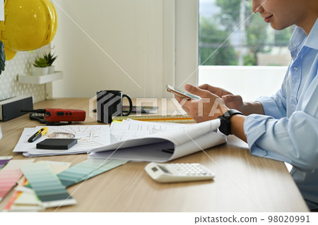 Architects man using mobile phone, working on design of a construction plan at creative workplace Architects man using mobile phone, working on design of a construction plan at creative workplace 98020991
