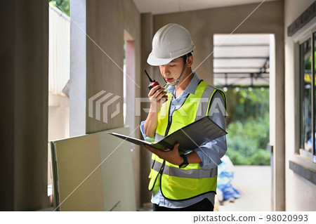 Civil engineer communicating with walkie talkie and working with blueprint at construction site 98020993