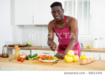 African american man with naked torso in an apron prepares vegetable salad in kitchen African american man with naked torso in an apron prepares vegetable salad in kitchen 98021405