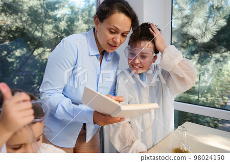 Multi-ethnic woman, a teacher wearing blue casual shirt, explaining chemistry lesson to smart kids in school science laboratory. Back to school. Knowledge. Education. Children's entertainment Multi-ethnic woman, a teacher wearing blue casual shirt, explaining chemistry lesson to smart kids in school science laboratory. Back to school. Knowledge. Education. Children's entertainment 98024150