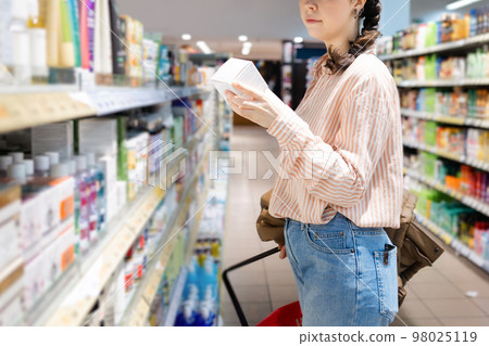 Close up of side view of Caucasian woman choosing cosmetic products in grocery store. Shelves with products in background. Concept of shopping and consumerism 98025119
