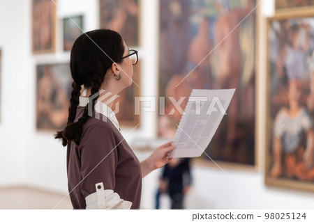 Side view of caucasian pretty young woman wearing glasses holds educational brochure. Defocused pictures in background. Concept of exhibition in gallery 98025124
