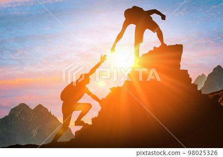 Man is giving helping hand. Silhouettes of people climbing on mountain at sunset. Help and assistance concept. Silhouettes of two people climbing on mountain and helping. 98025326