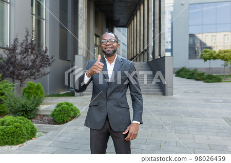 Portrait of a successful African American man. A businessman, a lawyer stands in front of an office center in a suit, looks at the camera, points a super finger, smiles. 98026459