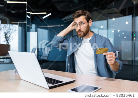 Bankruptcy, financial problems, unsuccessful online shopping. Shocked young man sitting in the office, holding a credit card, looking at a laptop. Checks the account, scratches the back of the head. 98026461