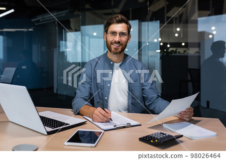 Portrait of a young handsome man, freelancer, businessman, accountant. Sitting in the office, looking at the camera, smiling. Works on a laptop, with documents, fills in forms with a pen. Portrait of a young handsome man, freelancer, businessman, accountant. Sitting in the office, looking at the camera, smiling. Works on a laptop, with documents, fills in forms with a pen. 98026464