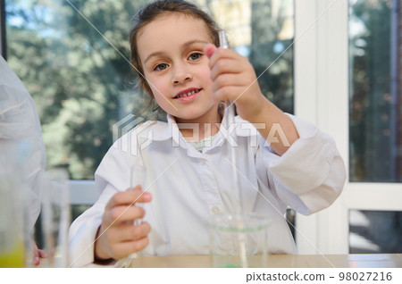 Close-up portrait of a Caucasian blue-eyed lovely little child girl in white lab coat, holding a test tube and a graduated pipette at chemistry class, in laboratory, cutely smiling looking at camera Close-up portrait of a Caucasian blue-eyed lovely little child girl in white lab coat, holding a test tube and a graduated pipette at chemistry class, in laboratory, cutely smiling looking at camera 98027216