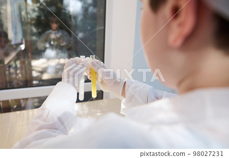 Rear view: selective focus on the test tubes with chemicals in gloved hands of a smart teenage schoolboy in white lab coat, conducting chemical scientific experiment in a chemistry laboratory Rear view: selective focus on the test tubes with chemicals in gloved hands of a smart teenage schoolboy in white lab coat, conducting chemical scientific experiment in a chemistry laboratory 98027231