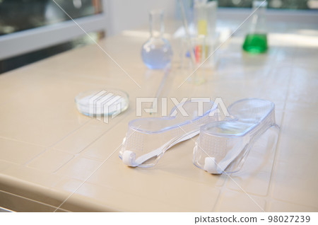 Selective focus on protective goggles - safety lab eyeglasses, eyewear on a plastic table, against the background of laboratory glassware Selective focus on protective goggles - safety lab eyeglasses, eyewear on a plastic table, against the background of laboratory glassware 98027239