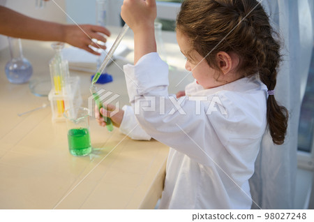 Side view of a Caucasian smart kid girl in white lab coat, using graduated pipette and measuring glassware, doing chemical experiment in school chemistry laboratory. Knowledge Education School concept Side view of a Caucasian smart kid girl in white lab coat, using graduated pipette and measuring glassware, doing chemical experiment in school chemistry laboratory. Knowledge Education School concept 98027248