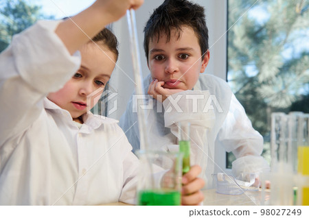 Caucasian smart teen schoolboy in white lab coat, watches a chemical reaction taking place in test tube and beaker, while conducting with his classmate an experiment in laboratory at chemistry class Caucasian smart teen schoolboy in white lab coat, watches a chemical reaction taking place in test tube and beaker, while conducting with his classmate an experiment in laboratory at chemistry class 98027249