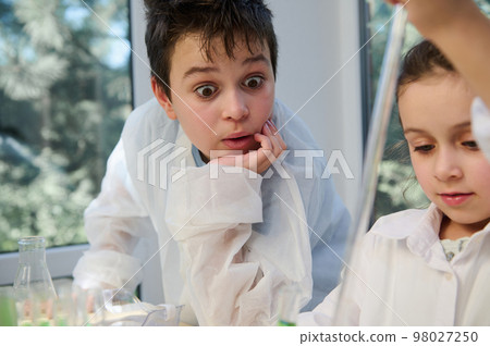 Amazed teenage boy in white lab coat, watching with stupefaction a chemical reaction taking place in the test tube in his classmate's hands, while conduct scientific experiment. Miracles of Chemistry Amazed teenage boy in white lab coat, watching with stupefaction a chemical reaction taking place in the test tube in his classmate's hands, while conduct scientific experiment. Miracles of Chemistry 98027250