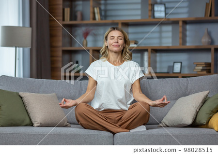 Senior woman at home resting meditating with eyes closed sitting on sofa in living room, housewife mature blonde in lotus pose smiling. 98028515