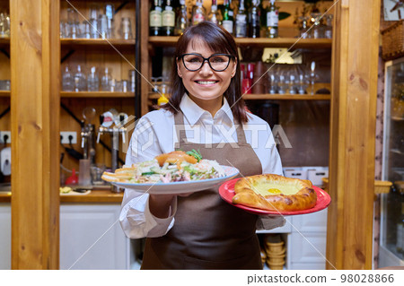 Middle aged female restaurant owner in apron with plates of cooked food Middle aged female restaurant owner in apron with plates of cooked food 98028866