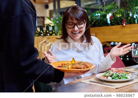 Woman sitting in restaurant, rejoices at plate of cooked food in hands of waiter 98028867