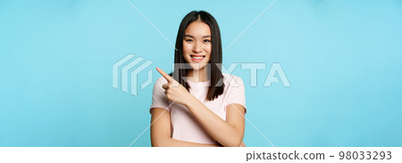 Smiling young asian woman 20 years old, pointing finger at upper right corner, showing promo banner, blue background Smiling young asian woman 20 years old, pointing finger at upper right corner, showing promo banner, blue background 98033293