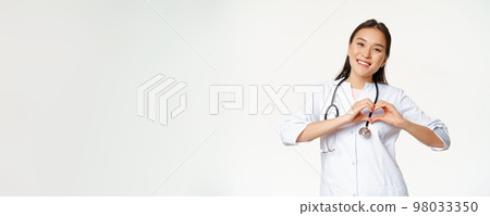Smiling asian female doctor care for her patients, shows heart gesture and looking happy, standing in medical clinic uniform, white background 98033350