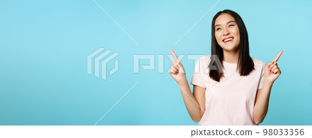 Smiling brunette asian woman, pointing sideways, showing both ways and laughing, choosing in store, standing over blue background 98033356
