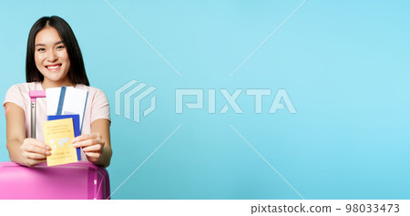 Happy korean woman, tourist with suitcare shows her health passport and tickets, covid-19 international vaccination certificate for travelling, going on trip, blue background 98033473