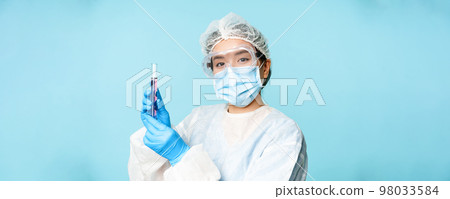 Asian nurse or lab worker in personal protective equipment, showing test sample tube, standing in medical face mask over blue background 98033584