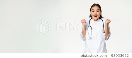 Enthusiastic female asian doctor celebrating, looking hopeful and happy at camera, triumphing, standing in medical uniform against white background 98033632